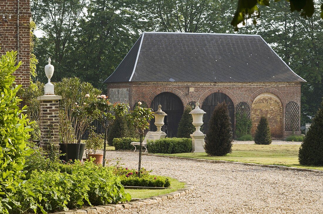 A gravel driveway leading to a country house with a view of the front garden with pedestals and a side house with rounded archways