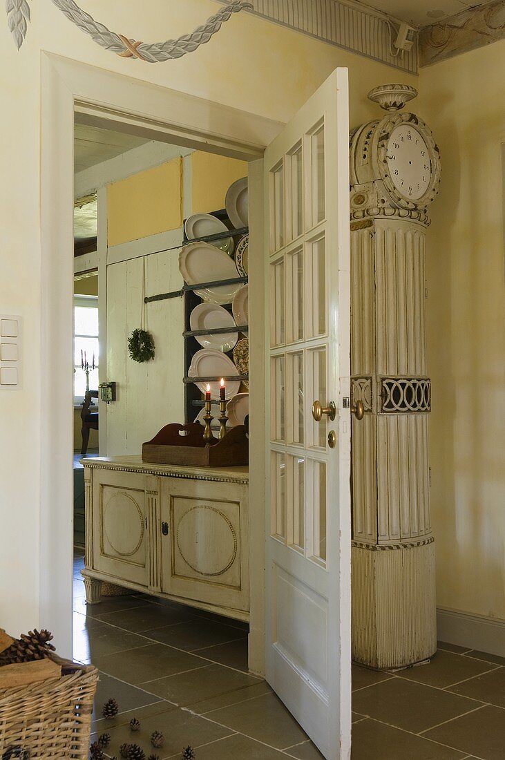 An antique grandfather clock standing next to an open door with a view of a plate rack in a dining room