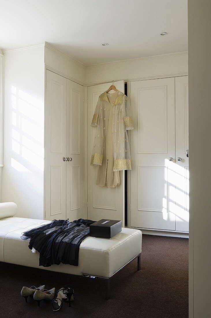 A built-in wardrobe in a dressing room with clothing on a white leather lounger