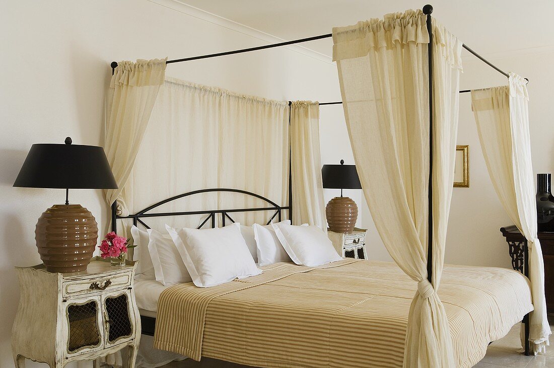 A metal four poster bed with a natural-coloured curtain and a vintage bedside table