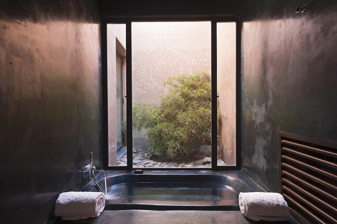 A concrete bathroom - a sun bathtub filled with water in front of a floor-to-ceiling window with a view of a courtyard