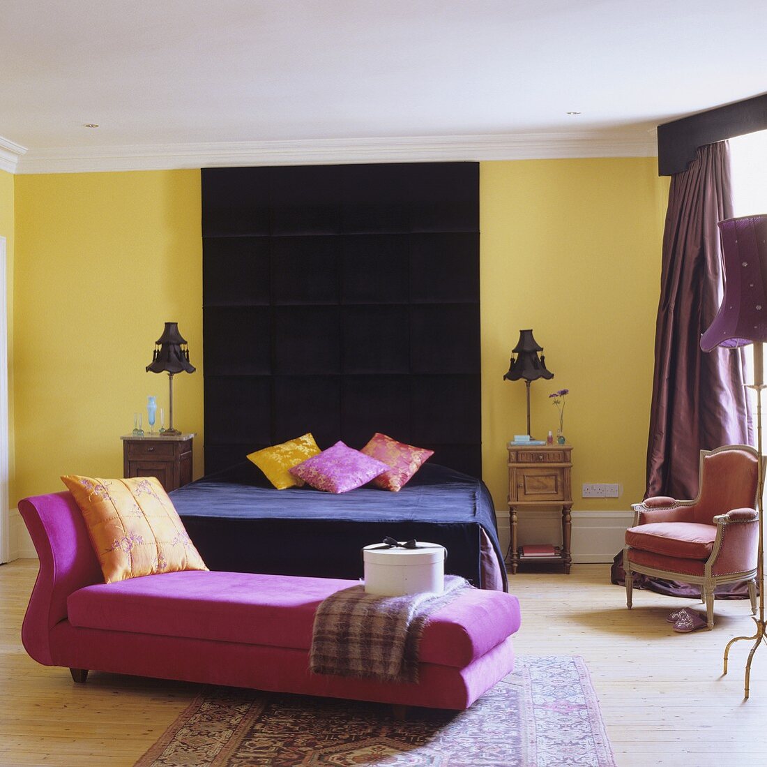 A colourful bedroom - a pink chaise longue with a black bed in the background with a ceiling-high headboard on the yellow wall