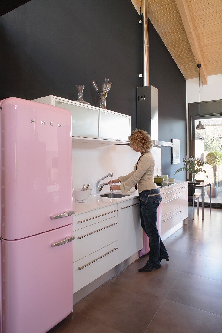 A kitchen with a 1950s-style fridge in front of a black wall and a woman at the sink