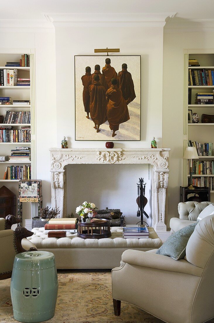 An upholstered coffee table in front of a fireplace with a white mantelpiece and a picture hanging above it