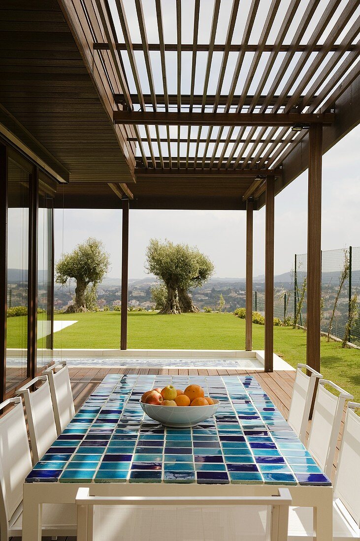 A table with a blue mosaic surface on a wooden terrace with a wooden slatted roof and a view of the garden