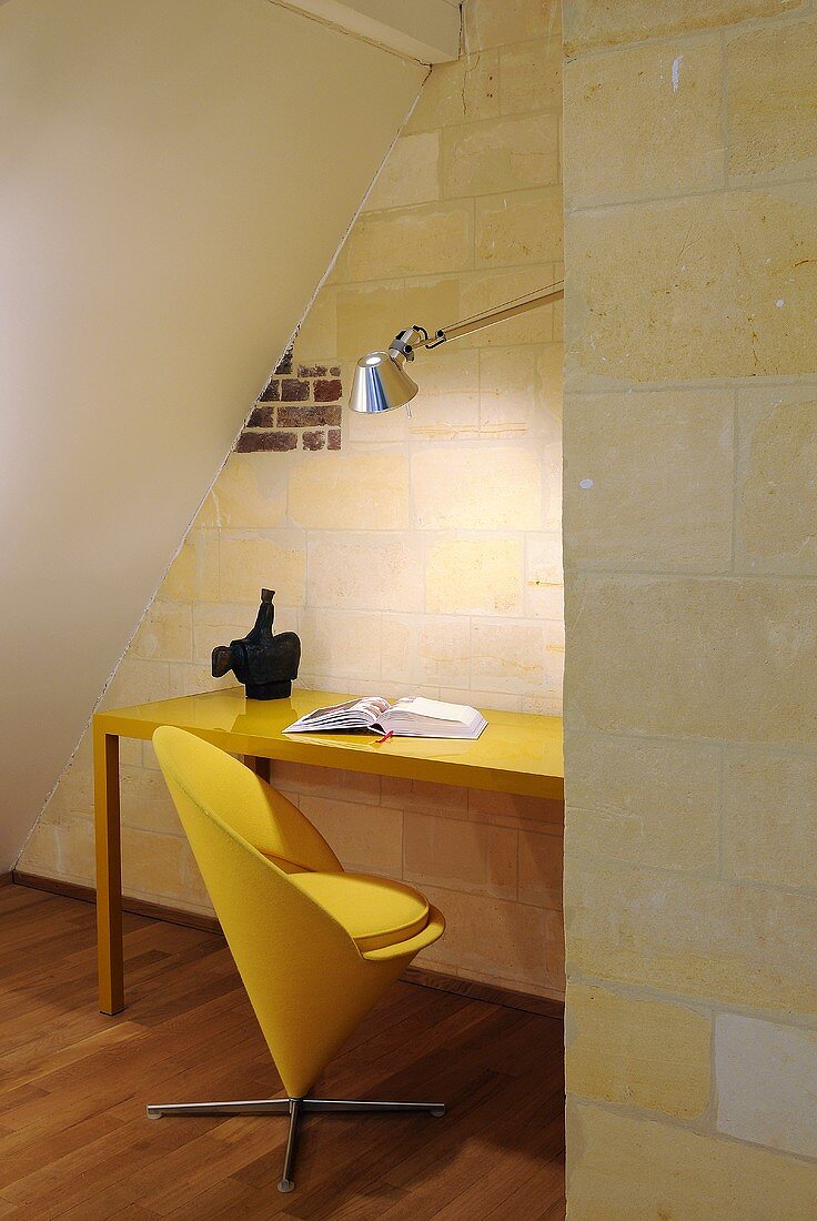 Minimalistische Arbeitsecke unter Dachschräge - gelber Klassikersessel vor Tisch und Natursteinwand