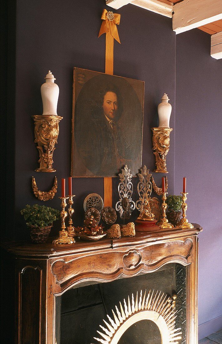 Holzgeschnitzter Kaminsims mit Kerzenständern und Goldkonsolen vor violetter Wand
