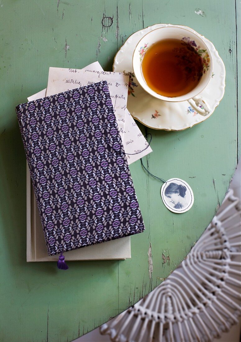 A cup of tea next to a book with a patterned cover on a green wooden surface
