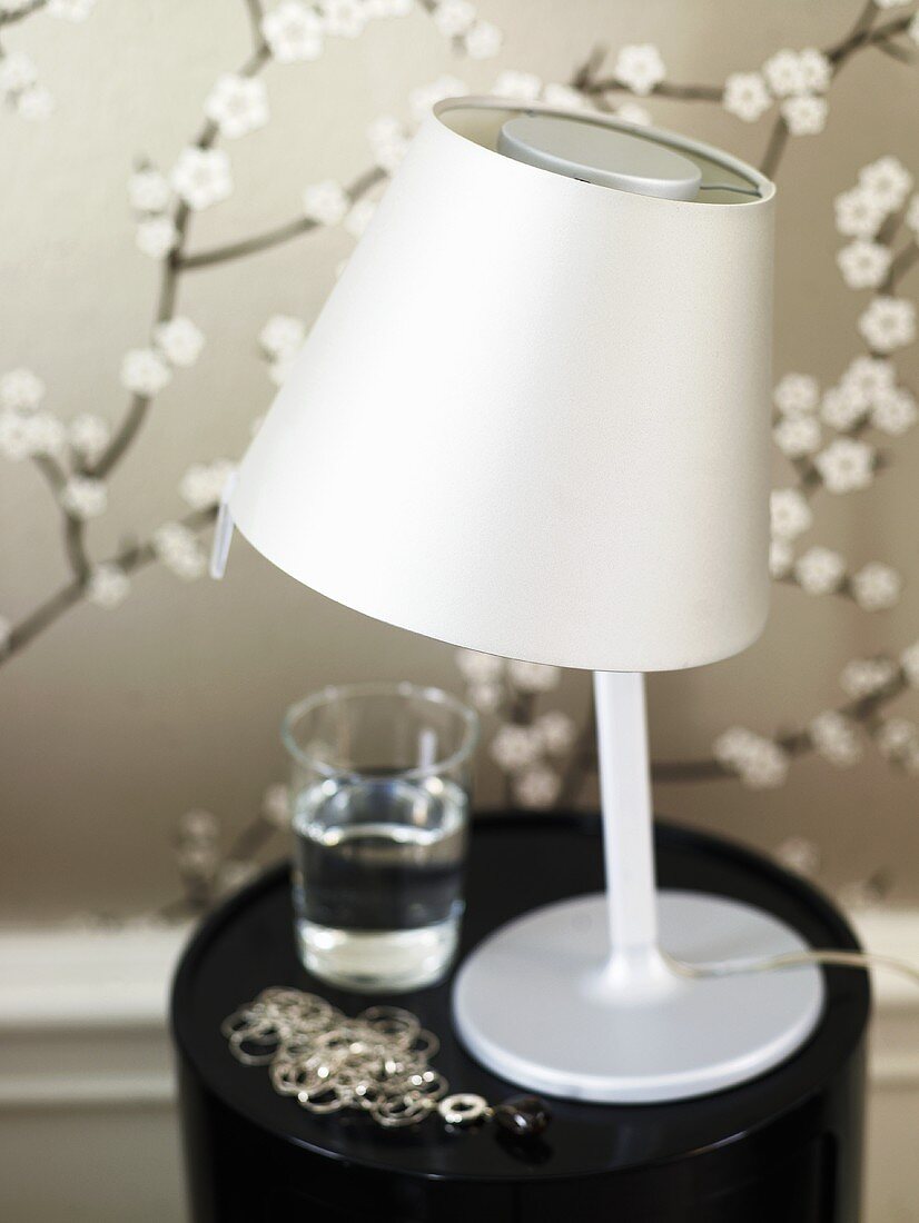 Table lamp with stainless steel shade on a black stool in front of patterned wallpaper