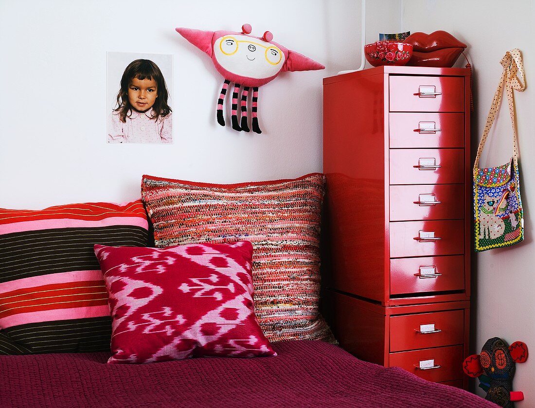 Kinderzimmerecke mit bunten Kissen auf Bett und rotem Metallcontainer mit Schubladen