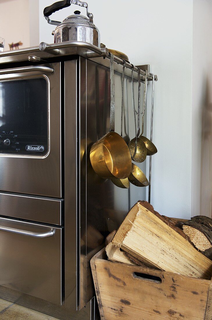 Kupfer Kochgeschirr an Metallgriff eines Küchenofens aus Edelstahl und Holzscheiten in Holzkiste