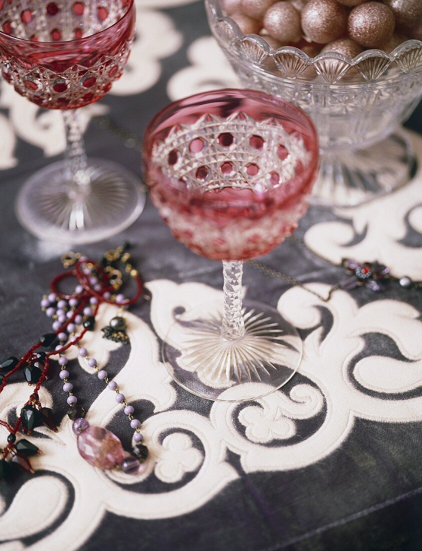 Rote Kristallweingläser und Perlenketten auf Tischdecke mit Ornamentmuster