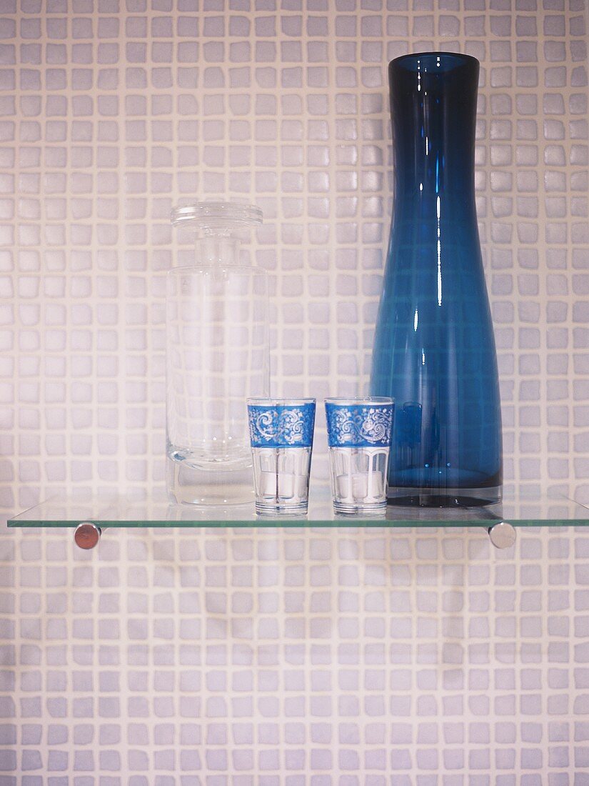 A detail of a modern bathroom, a glass shelf with blue glass vase, decorative glasses, mosaic tiles,