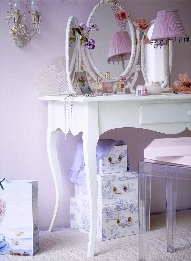 A detail of a pink bedroom showing a white painted dressing table, three sided vanity mirror, lamp, storage boxes,