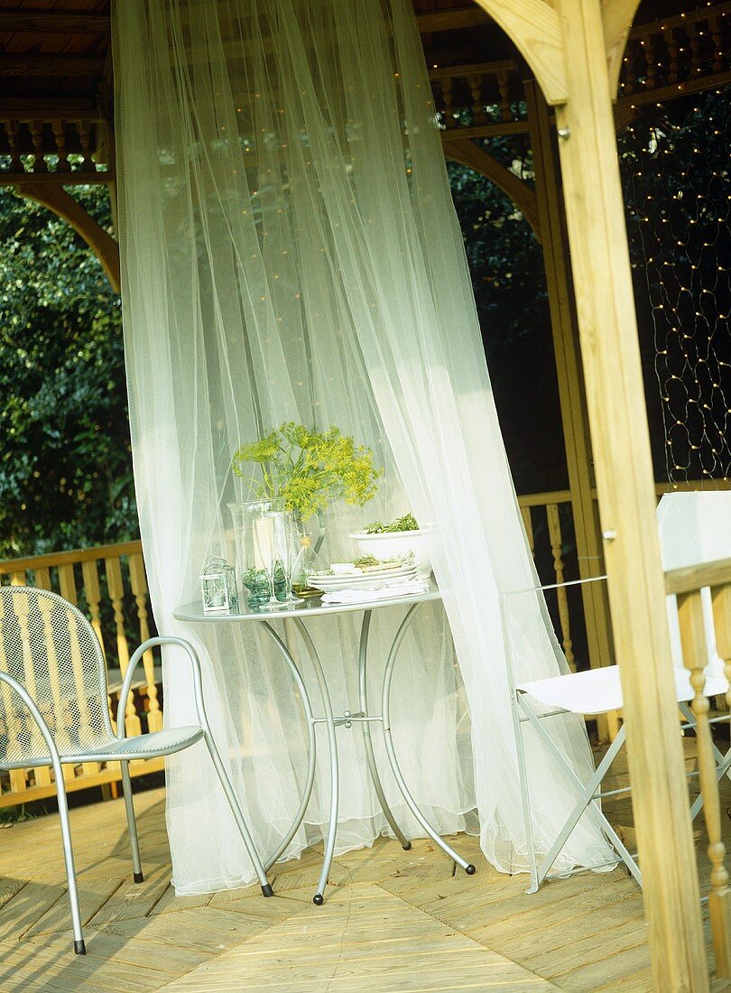 Metal table, draped with white sheer curtain, set for meal in gazebo.