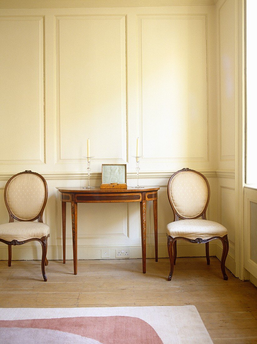 Antiker Wandtisch und Stühle mit gepolsterter Rückenlehne und Sitzfläche im Rokokostil vor weisser Holzvertäfelung