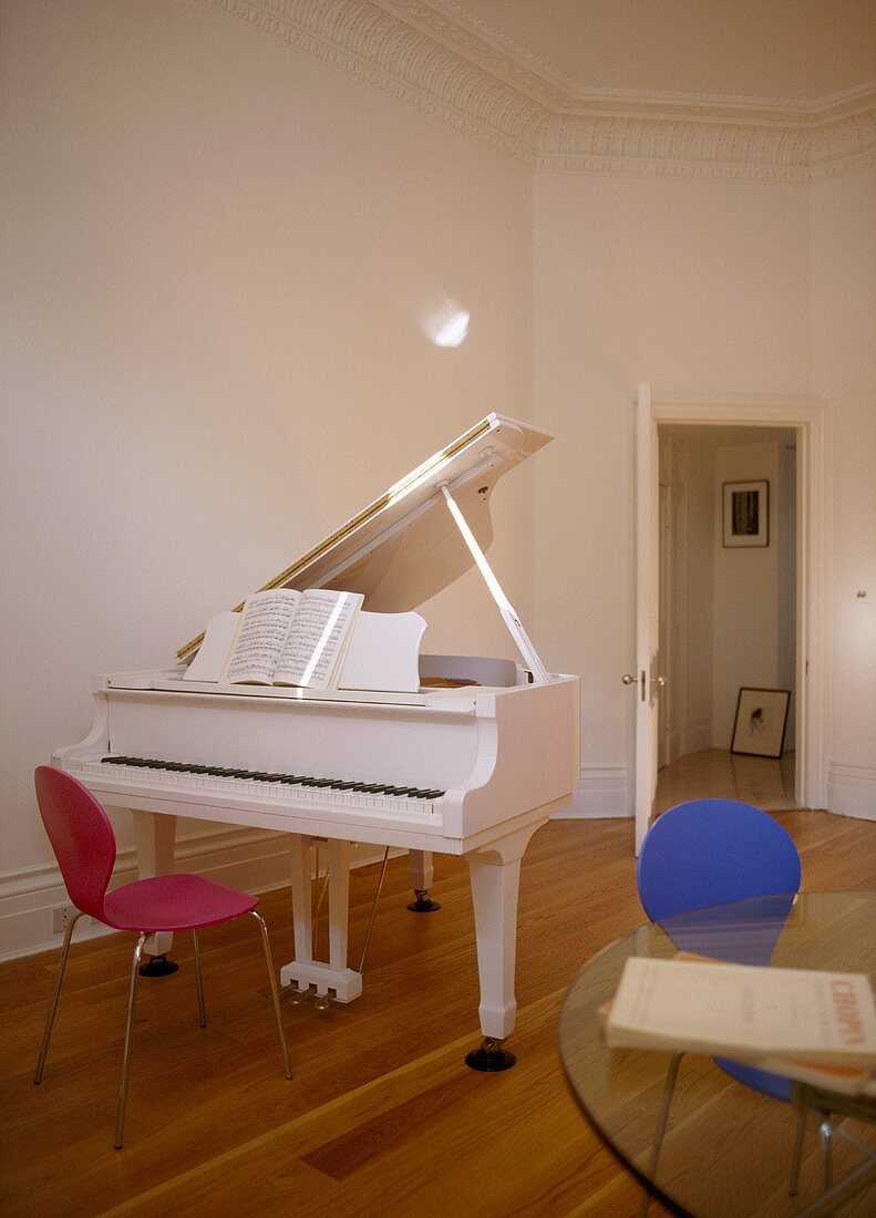 A detail of a music room, a white piano, red retro chair, wood floor, glass table, door open,