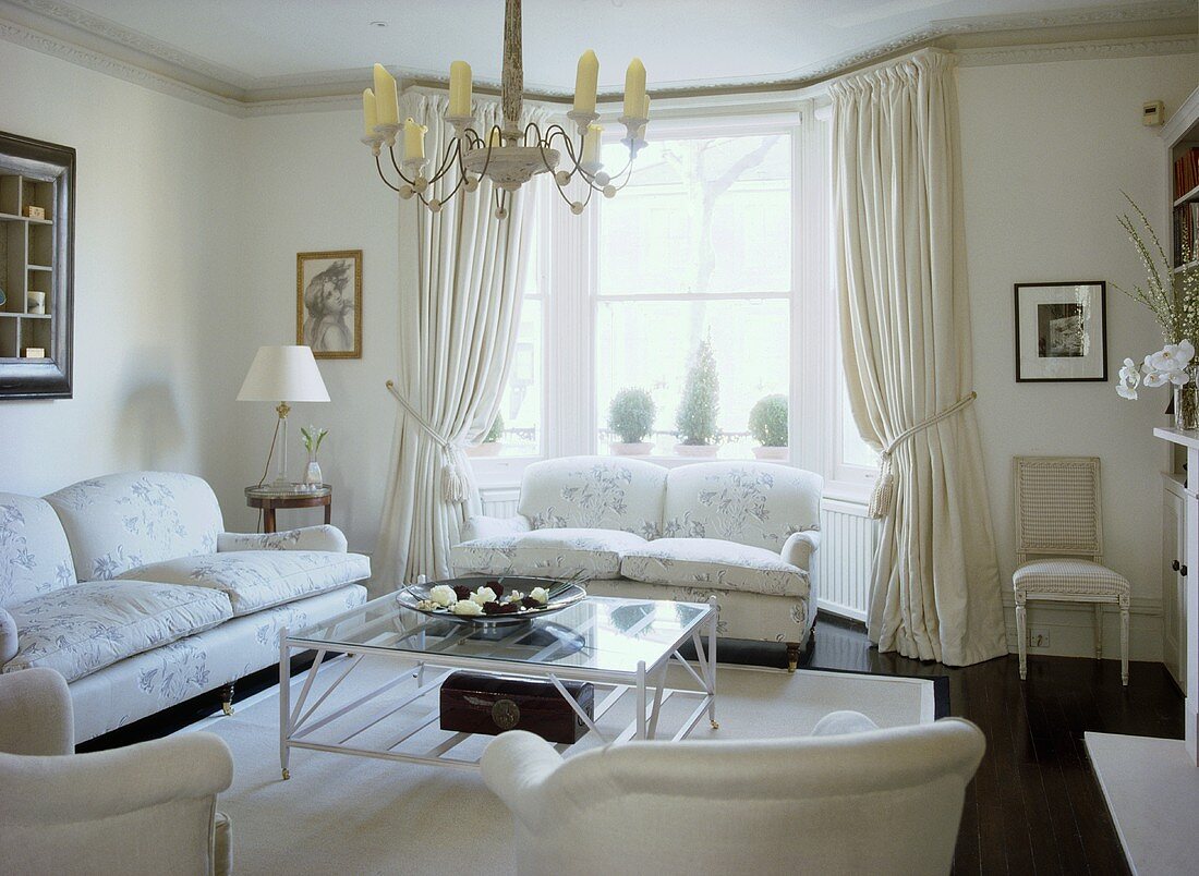 A traditional, neutral sitting room with upholstered sofas, glass coffee table, curtains, chandelier