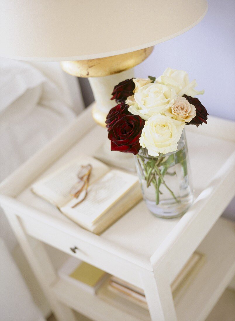 A detail of a modern bedroom, showing a white painted bedside table, lamp and roses in a glass