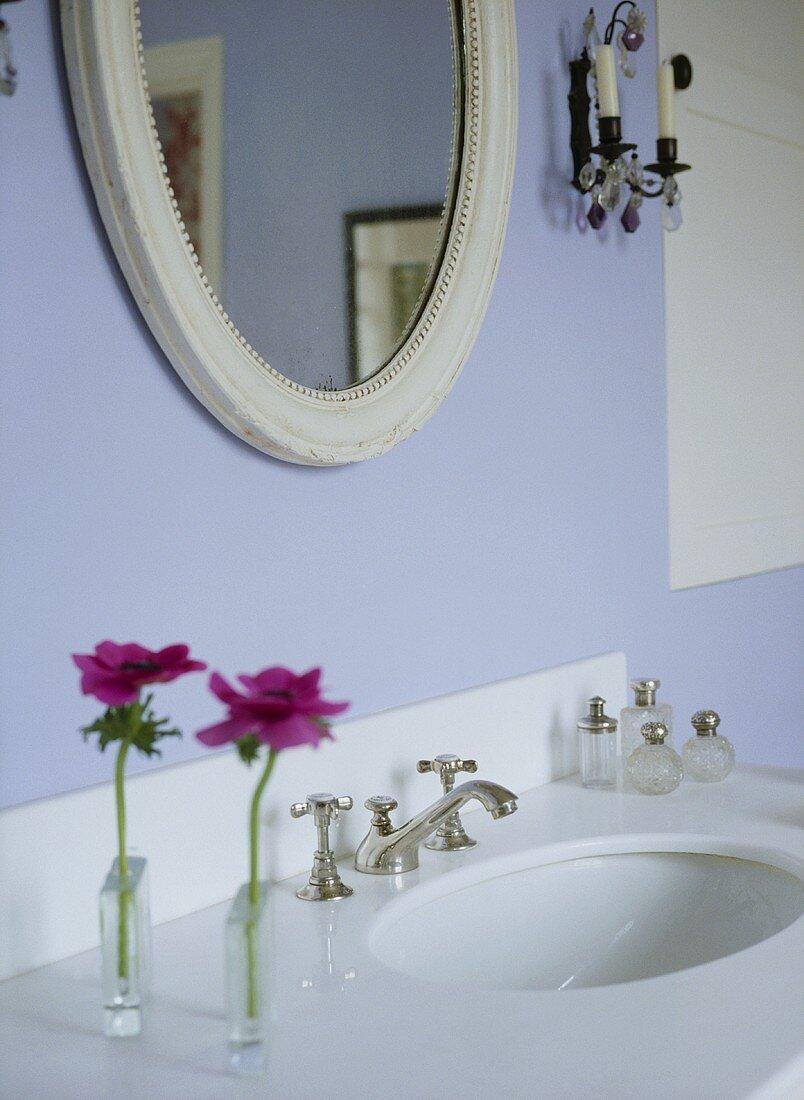 A detail of a modern, pale blue bathroom showing washbasin set in unit, oval mirror, glass perfume bottles, styled