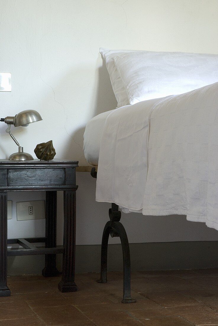A bedside table next to a high bed with white bedclothes