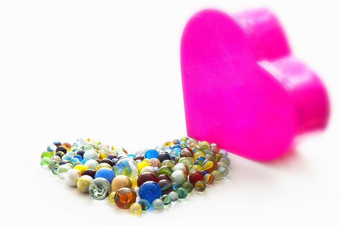 Colourful glass marbles in the shape of a heart next to a pink heart-shaped chocolate box