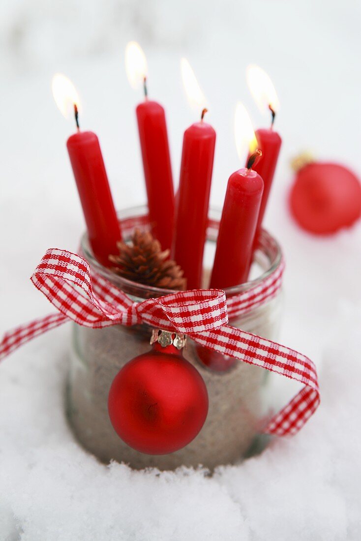 Rote Kerzen in einem Einmachglas mit Geschenkband und Weihnachtskugel im Schnee