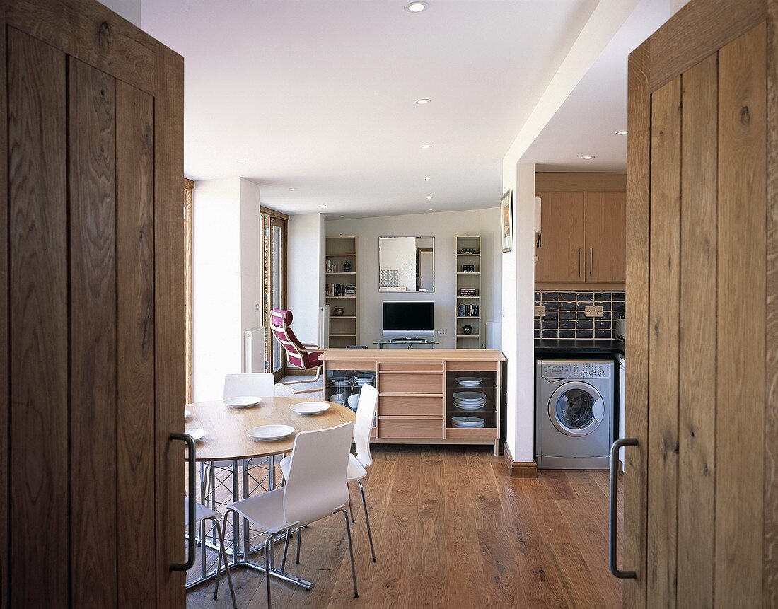 View through an open wooden double door to a dining area with white chair in front of an open kitchen-living room