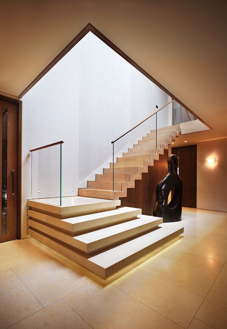 Elegant entry area with angled, stone staircase and glass banister beside a modern sculpture