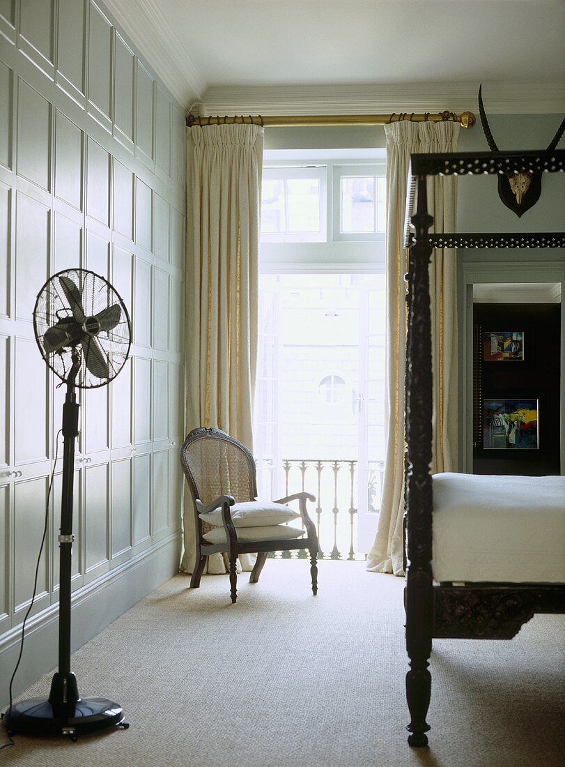 A traditional bedroom with painted panelling, carved wooden four poster bed, cream curtains, armchair, floor fan
