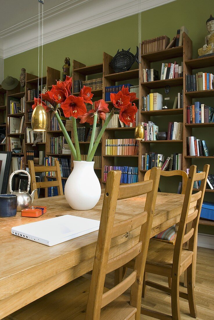 Red amaryllis in a white vase on a wooden dining table with a bookshelf in the background