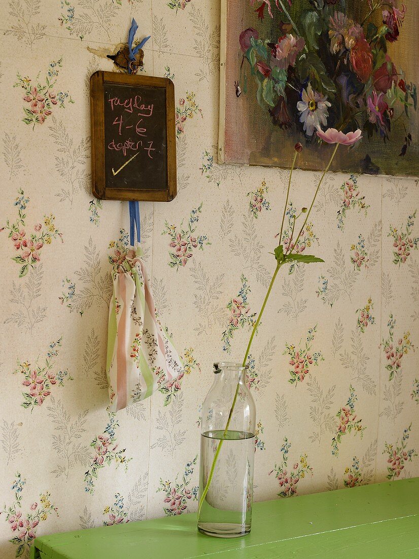 A flower in a glass bottle in front of a wall hung with floral paper