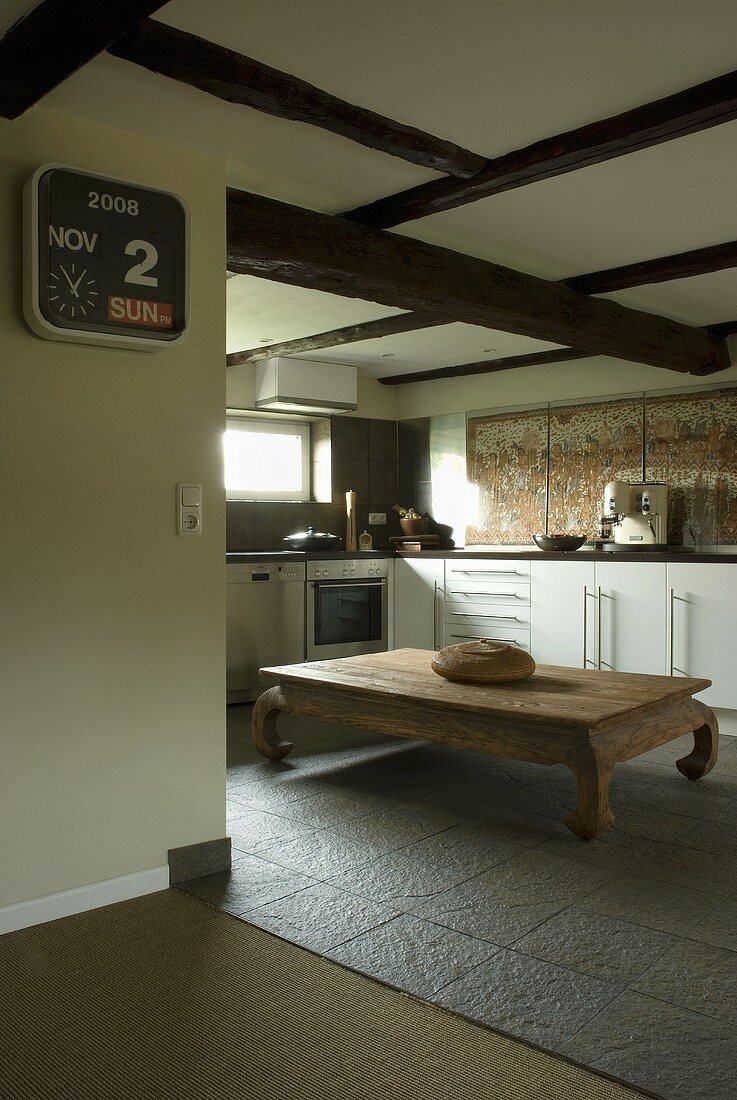 Offene Küche mit weissen Schrankfronten unter Holzbalkendecke und rustikalem Holztisch