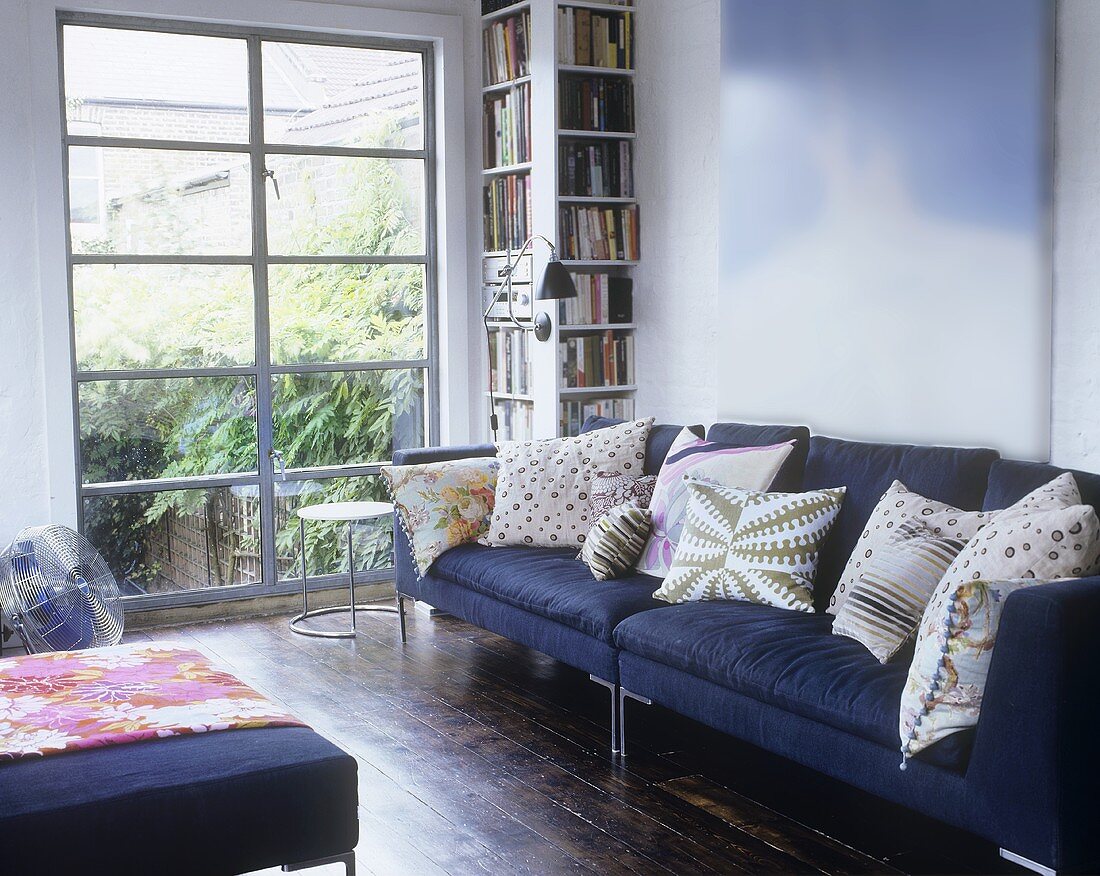 Violettes Sofa mit Kissen neben Bücherregal und raumhohem Fenster