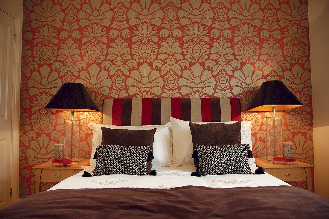 Doppelbett mit Nachttischen und Nachttischlampen vor rot-gold gemusterter Tapete