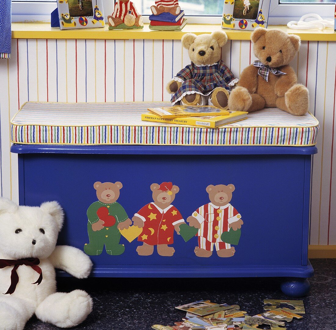 Blaue Holztruhe mit aufgemalten Teddybären und einem gestreiften Sitzkissen, darauf sitzen Teddybären