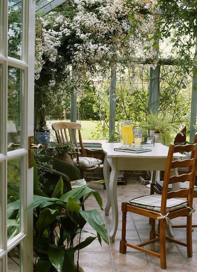 Offene Tür zu einem Wintergarten mit antiken, hölzernen Stühlen und weißem Tisch; darüber eine weissblühende Pflanze