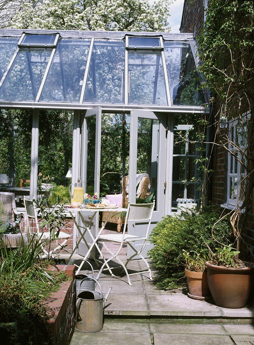 Terrasse mit weissen, metallenen Gartenmöbeln vor einem gläsernen Wintergarten