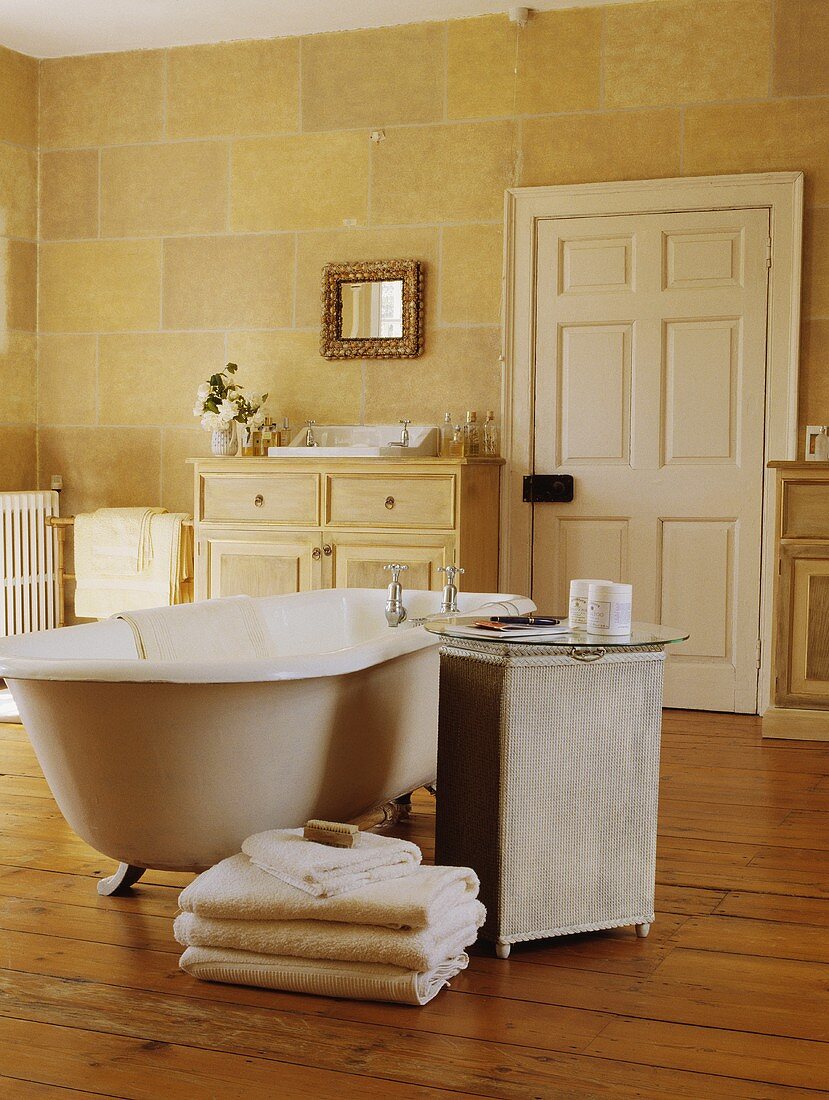 Badezimmer mit freistehender Clawfoot-Badewanne, einem Lloyd Loom Tisch mit Glasplatte, einem cremefarbenen Handtuchstapel und mit aufgemaltem Steinwand-Effekt an den Wänden