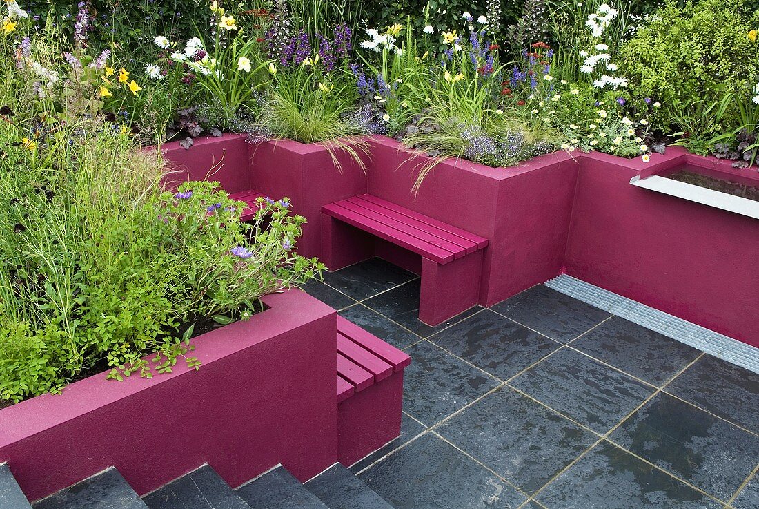 Terrasse mit grauen Fliesen, die pinkfarbenen Wände dienen als Abgrenzung von den Blumenbeeten