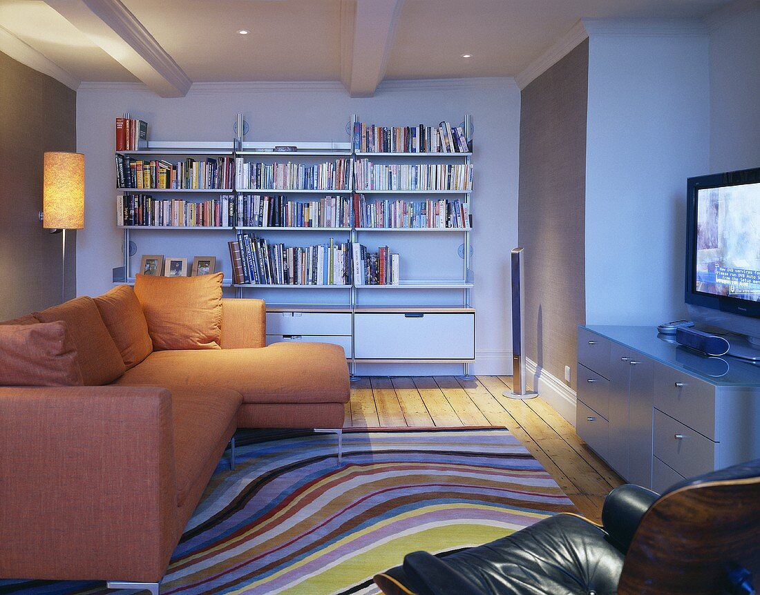 Offener Wohnraum mit orangefarbenem Sofa übereck vor Bücherregal und Teppich mit geschwungenen Streifen