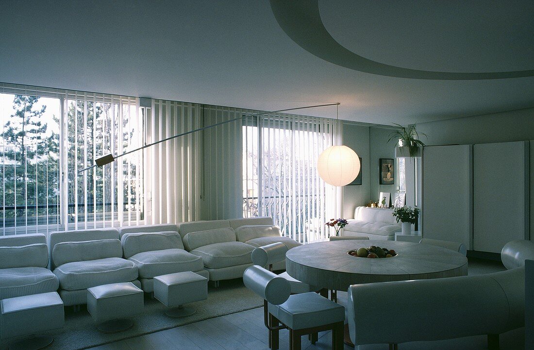 Weisses Sofa vor dem Fenster und runder Steintisch mit weissen Leder-Sitzbänken in einem offenen Wohn- und Esszimmer