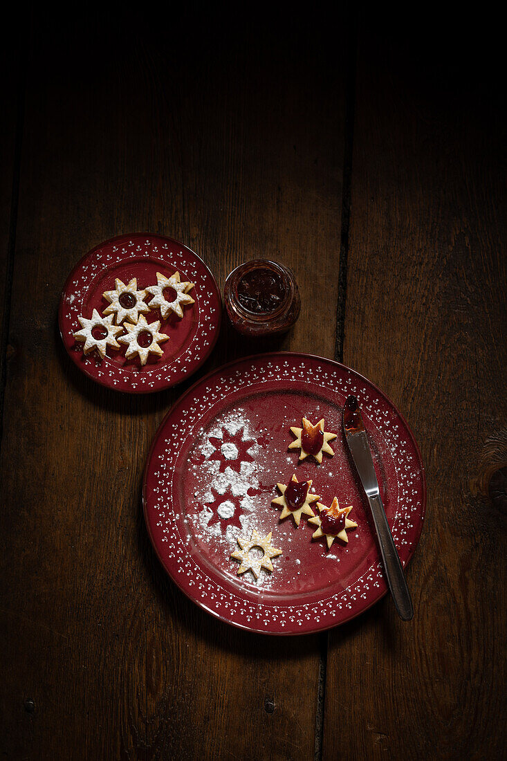  Christmas cookies (Spitzbuben) with jam, knife and on red plates, on wooden background  