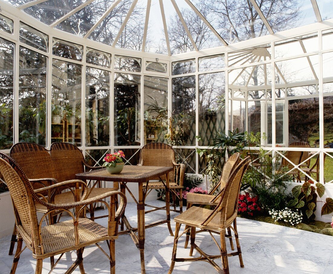 Korbstühle mit passendem Tisch in weißem, mit Beeten bepflanztem Wintergarten und Ausblick in den Garten