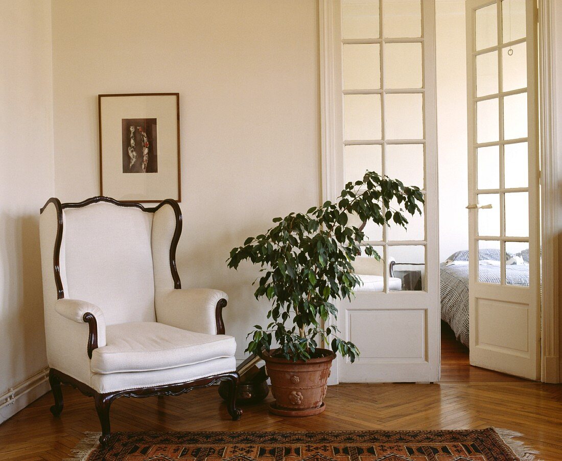 Vorraum mit weißem, antikem Ohrensessel vor kubistischer Malerei und Blick durch französische Fenstertüren in Schlafzimmer