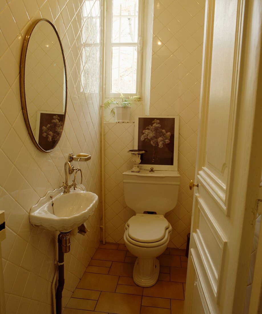 Blick durch offene Tür auf kleines, weisses WC mit Toilette und Waschbecken im antiken Stil zu ovalem Spiegel