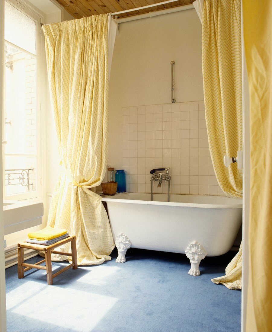 Lange, gelbe Vorhänge vor freistehender Badewanne mit Klauenfüssen auf blauem Teppich in traditionellem Bad