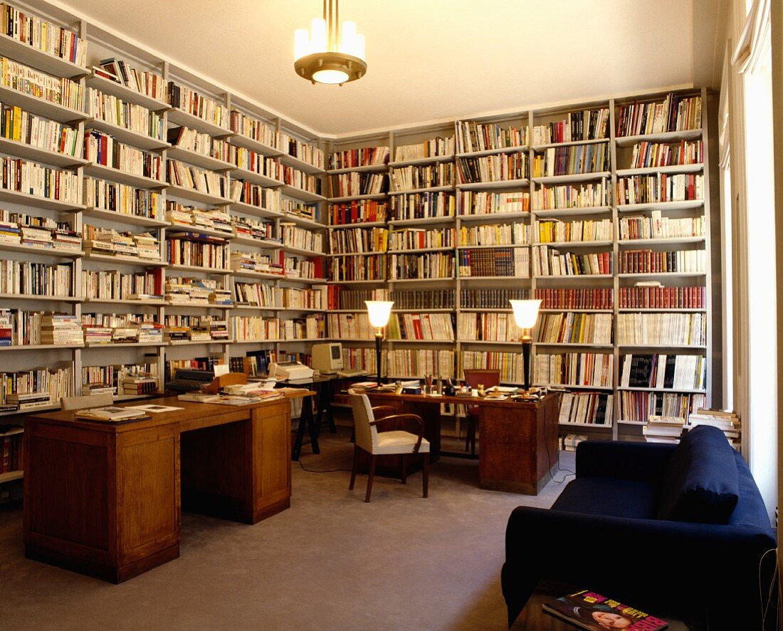 Grosse, alte Schreibtische und modernes, blaues Sofa in Bibliothek mit raumhohen, weissen Bücherregalen