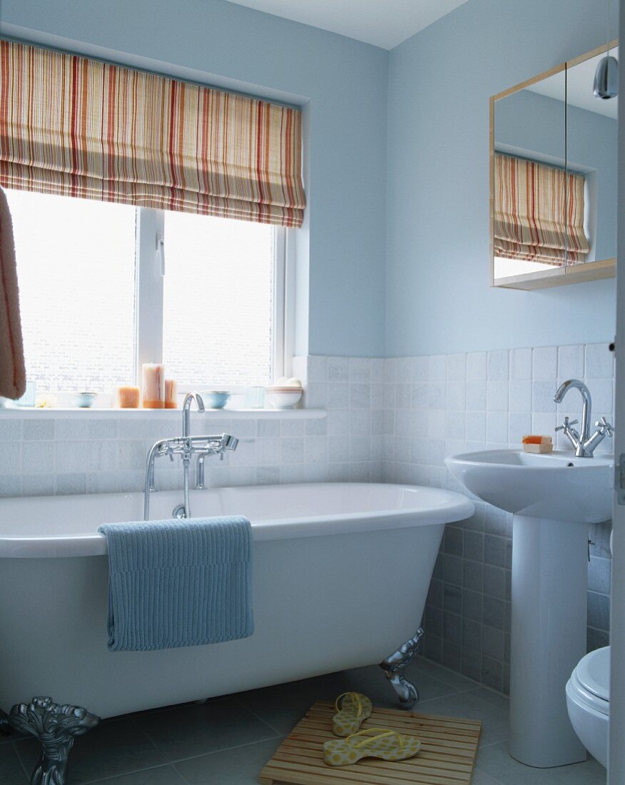 Naturfarben gestreifte Jalousie neben pastellblauer Wand und Klauenfüsse an freistehender Badewanne