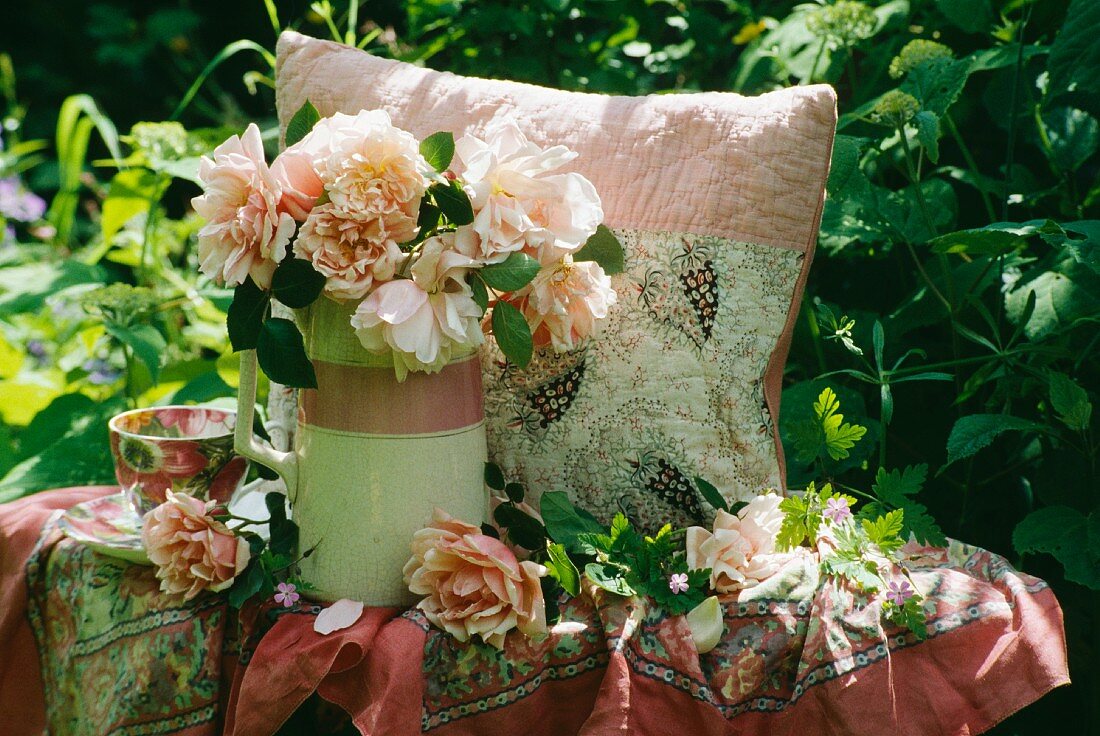 Dekoration roséfarbener Rosen, Krug und Blumentasse auf rosé gemustertem Stoff und Kissen im Grünen